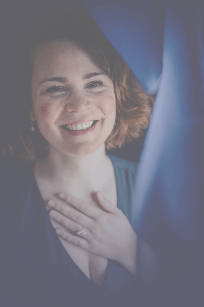 Frau mit dunkelblauem Kleid schaut hinter blauem Vorhang hervor, lachend und mit einer Hand auf dem Herzen. Sie lädt ein tiefzutauchen in Seelenbalsam-Sessions und Seelenbalsam-Kreisen.